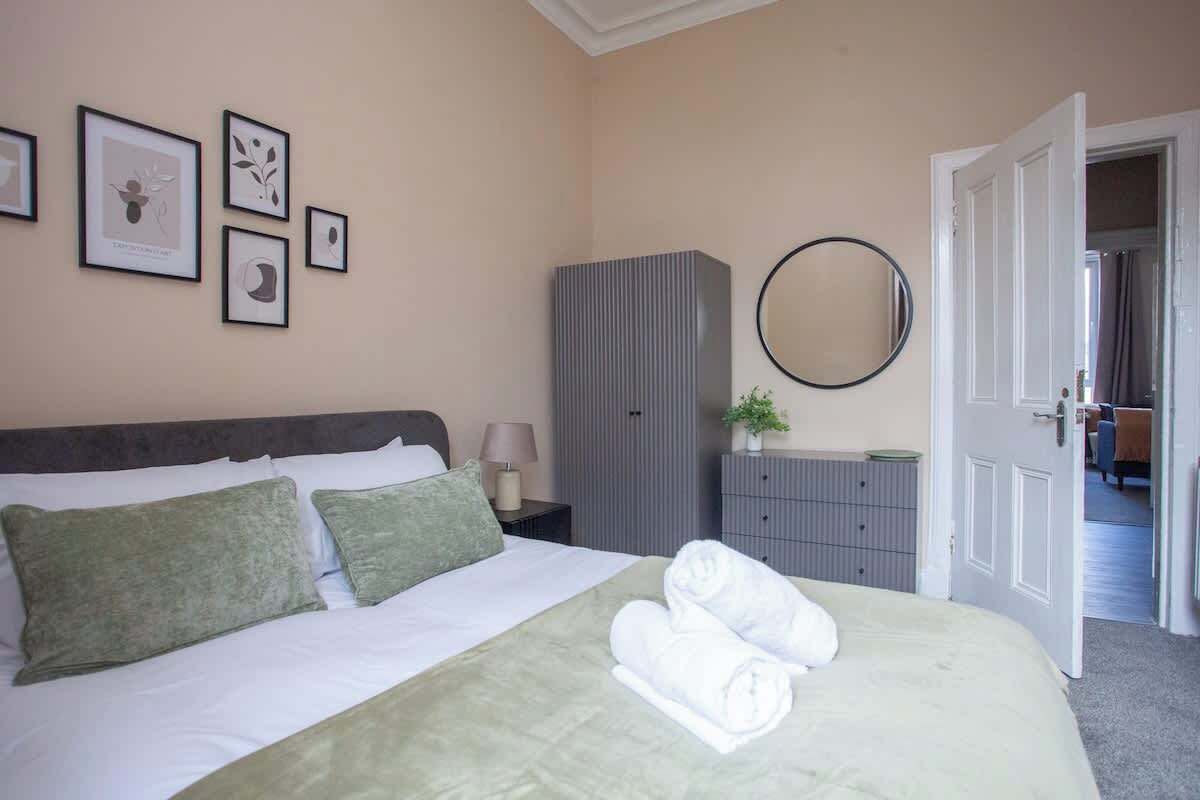 A neatly made bed with pillows, a grey wardrobe, a round mirror, and framed artwork on the wall.