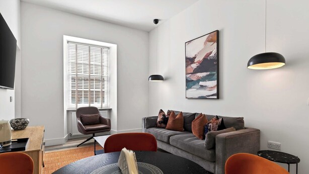 A modern living room with a grey sofa, orange chairs, a dining table with a placemat, a black pendant light, and a painting on the wall. - Modern City Apartment Just off Buchanan St (Glasgow City)