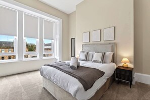 Une chambre avec un grand lit, une table de chevet avec une lampe et une vue dégagée sur l’extérieur grâce à de grandes fenêtres.