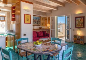 A dining area with a round table, chairs, and a bowl of fruit. There is a red sofa and a wooden cabinet with shelves. - Sky Rooftop Ortigia (Syracuse)