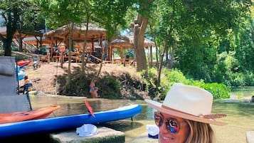 A person seated at a table with a drink, surrounded by a natural setting with trees and a river.