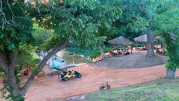 A dirt path leading to a water feature with a small bridge, surrounded by trees and outdoor seating areas.