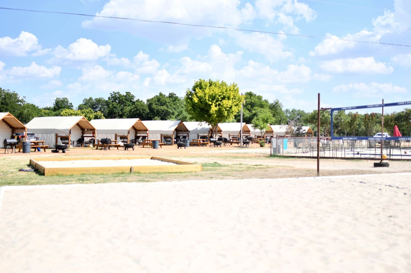 A row of tents with picnic tables and barbecue grills, set against a backdrop of trees and a clear sky.