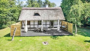 Ein Haus mit einem strohgedeckten Dach, einer Holzterrasse, einem Tisch mit Stühlen und einem Fenster.