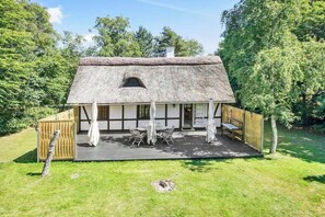 Ein Haus mit einem strohgedeckten Dach, einer Holzterrasse, einem Tisch mit Stühlen und einem Fenster.