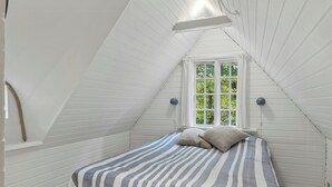Ein kleiner Raum mit geneigtem Dach, einem Bett, einem Fenster und einer Wandlampe.