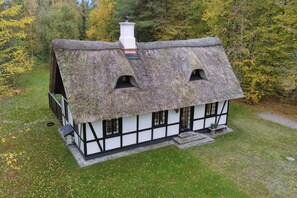 Una casa tradicional amb teulada de palla i façana blanca amb adorns negres, envoltada d'un jardí herbaci i arbres.