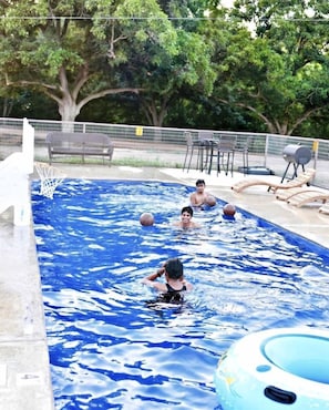 Una piscina con aro de baloncesto, sillas de descanso y un área cercada con árboles.