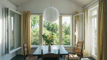 Une pièce avec une table et des chaises en bois, un vase rempli de fleurs et de grandes fenêtres avec des rideaux.