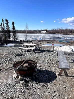 A camping area with picnic tables, a fire pit, and a view of a frozen lake and forest.