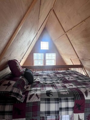 Ein gemütliches Schlafzimmer im Dachgeschoss mit schrägen Wänden, einem Fenster mit Aussicht und einem Bett mit kariertem Überwurf.