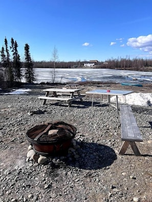 A camping area with picnic tables, a fire pit, and a view of a frozen lake and forest.