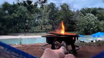 Uma pessoa relaxando perto de uma fogueira, rodeada por árvores e um corpo de água.