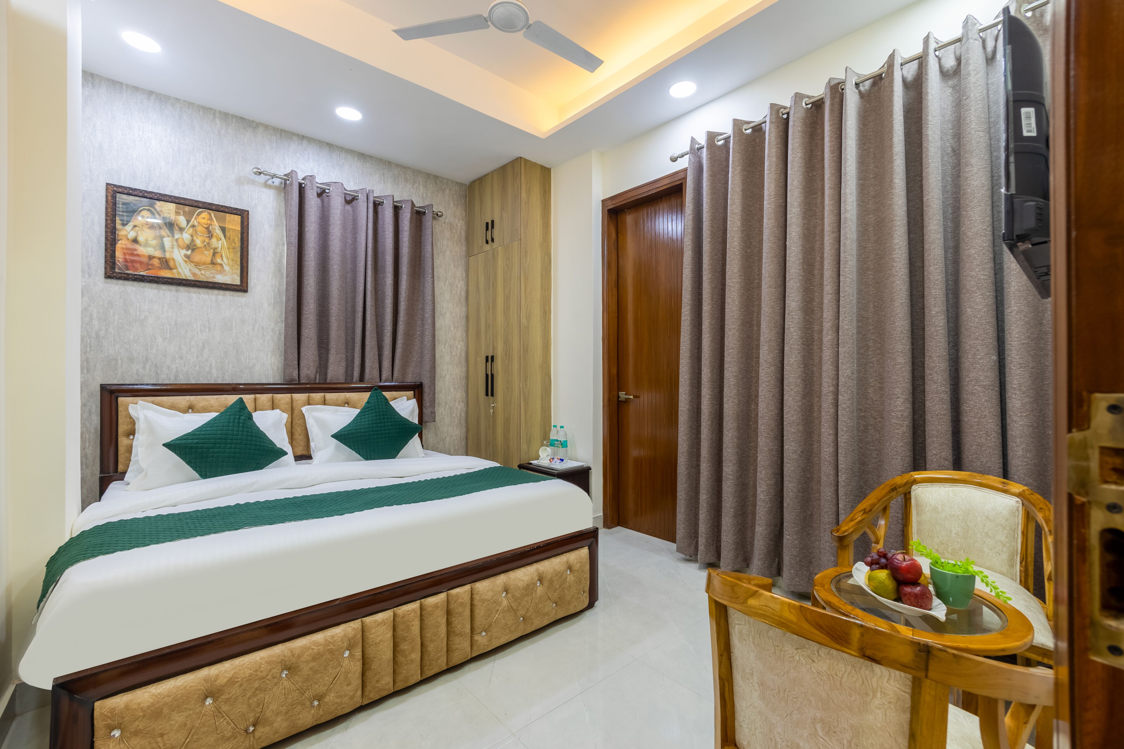 A hotel room with a bed, a nightstand, a chair with a tray table, curtains, a ceiling fan, and a wall-mounted TV.