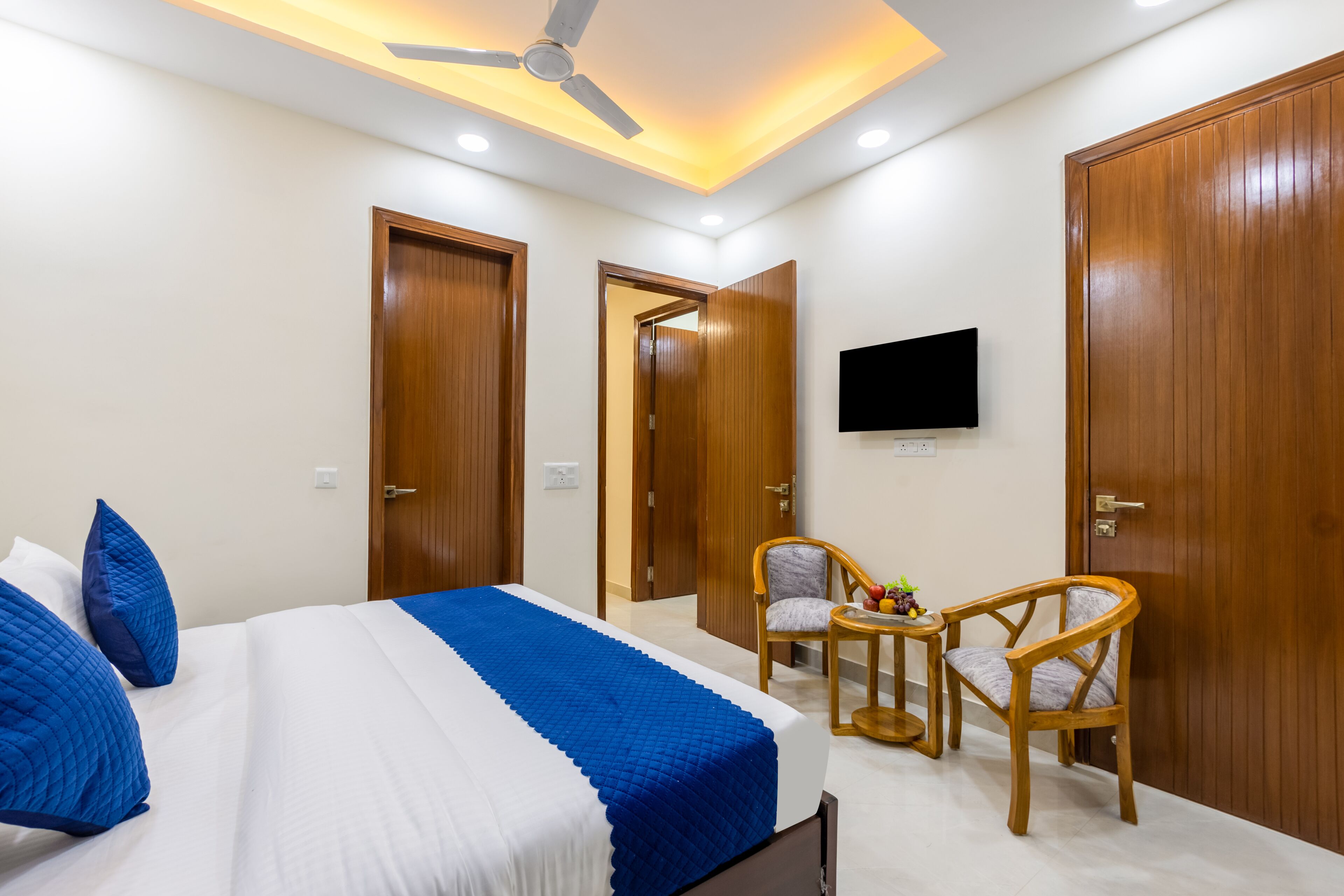 A hotel room with a bed, two wooden doors, a ceiling fan, a TV mounted on the wall, and two wooden chairs.