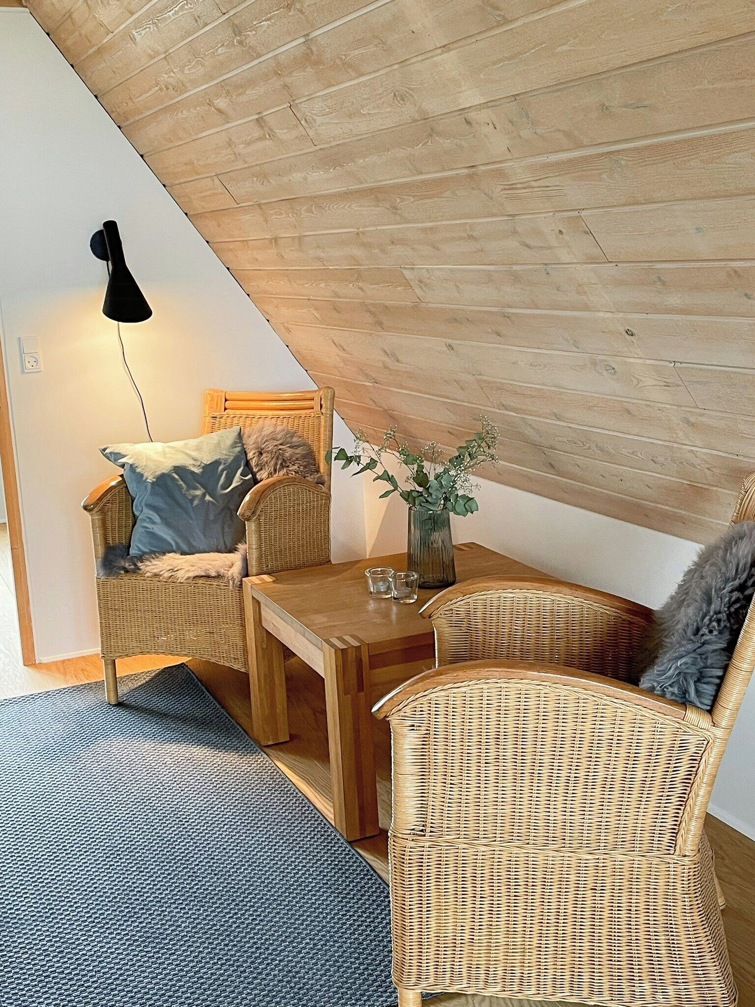 A cozy loft bedroom with a sloped ceiling, a wooden dining table, wicker furniture, and a small vase with flowers.