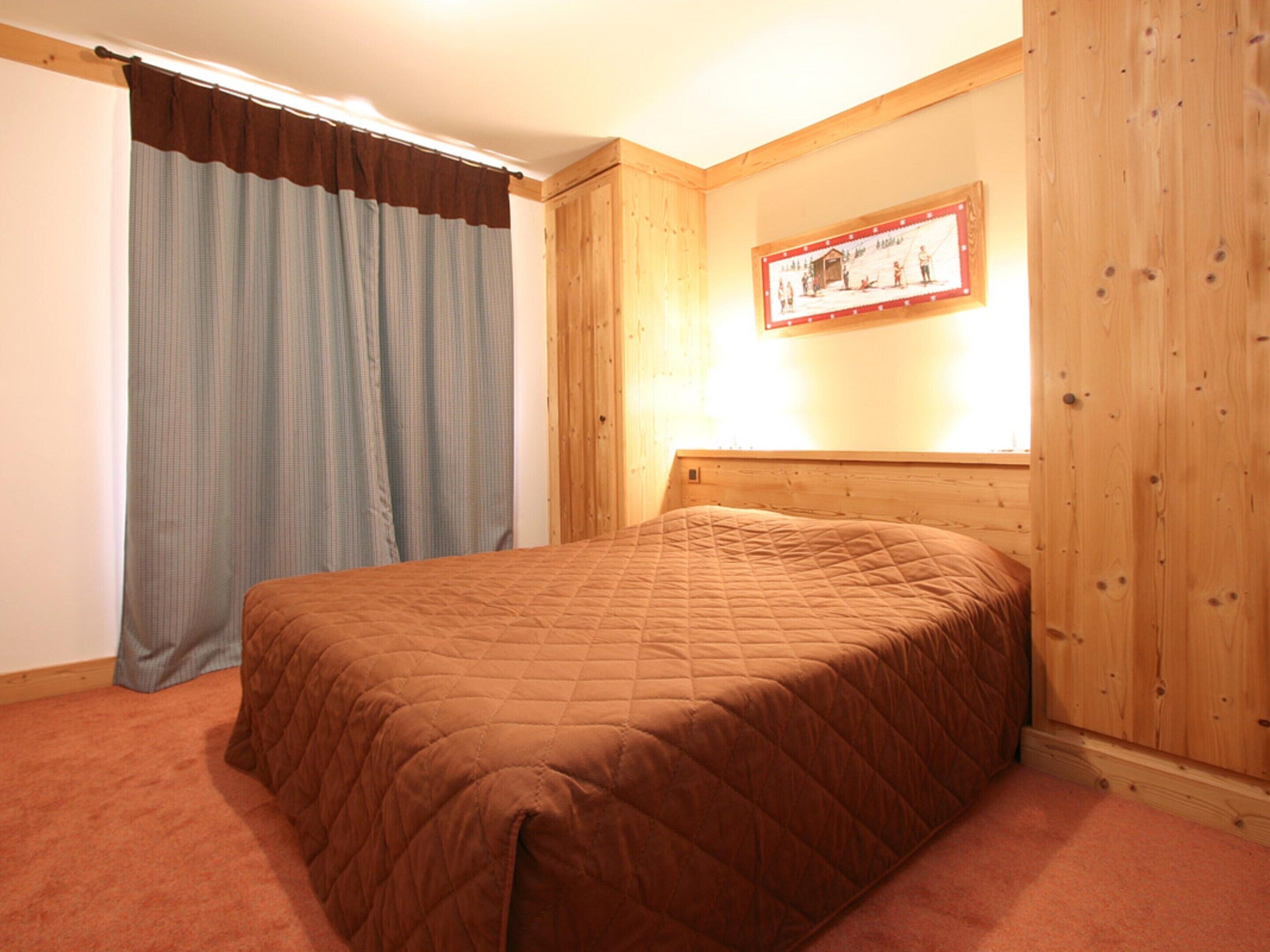Un dormitorio con cabecera de madera, cama con colcha marrón, ventana con cortinas e imagen enmarcada en la pared.