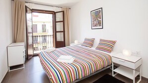 Quarto com cama, mesa de cabeceira, janela com cortinas e porta que dá para uma varanda.
