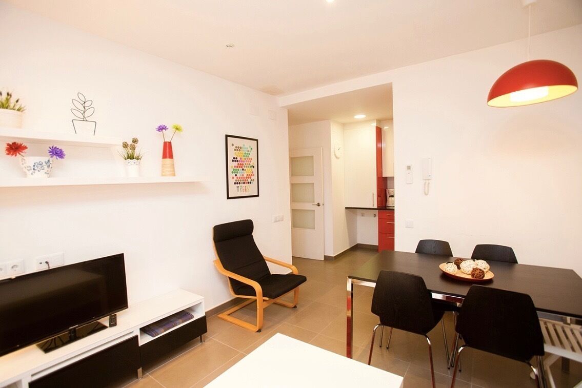 A modern living room with a flat-screen TV, a black chair, a dining table with chairs, and a red pendant light.
