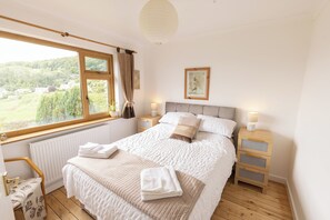 Un dormitorio con un ventanal grande, una cama con ropa de cama blanca, una mesita de noche con lámpara y suelo de madera.