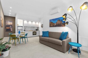 Een moderne woonkamer met een bank, een eettafel met stoelen, een keuken en een groot raam.