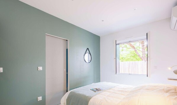 Une chambre à coucher avec un lit, une fenêtre donnant sur des arbres, une porte et un luminaire fixé au mur.