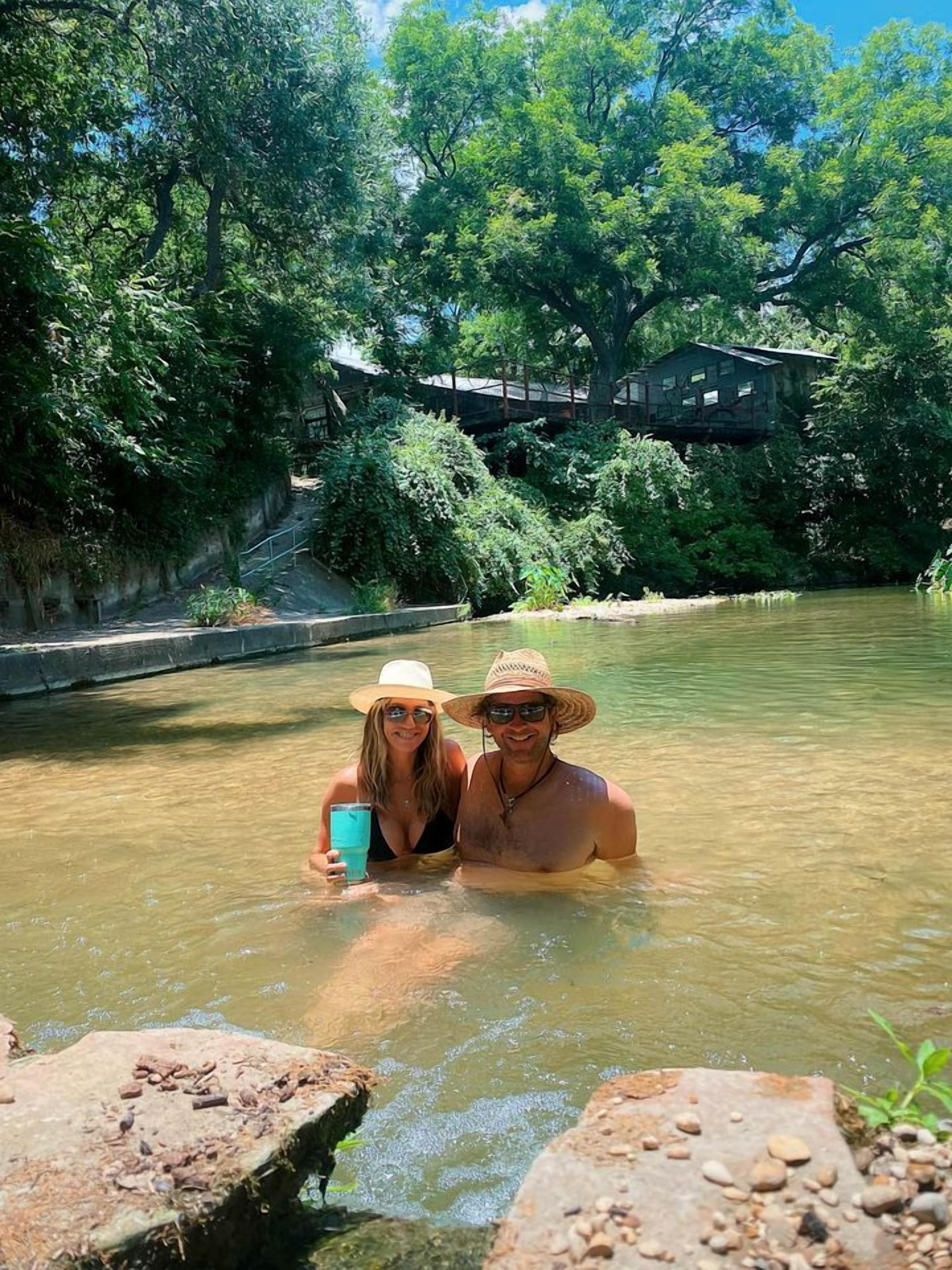 Una pareja en un entorno natural con un cuerpo de agua, rodeada de vegetación y una estructura elevada sobre pilares.