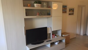 A modern living room with a flat-screen TV, a lamp, and a shelf with decorative items. - 'Prestige Apartment' with Shared Pool and Private Garden (Marina di Bibbona)