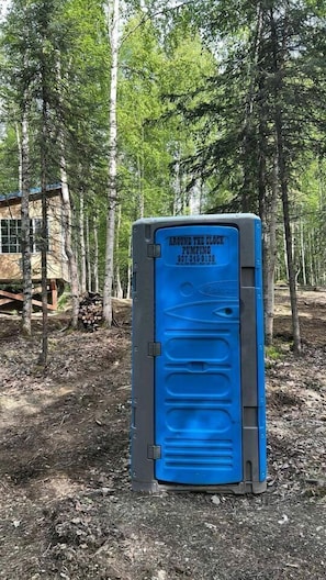 A portable toilet with a blue door, labeled 'Around the Clock Pumping,' in a wooded area. - Relaxing Mini Cabin Hideaway with Campground Amenities, Play Area & Firepit in Houston, Alaska (Houston)