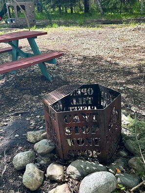 A metal fire pit with the words 'CAMPING CAN'T STOP WONDER' engraved on it, surrounded by rocks and a picnic table in a wooded area. - Relaxing Mini Cabin Hideaway with Campground Amenities, Play Area & Firepit in Houston, Alaska (Houston)