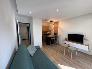 Ein modernes Wohnzimmer mit einer Couch, einem Fernseher, einem kleinen Essbereich und einer Küche im Hintergrund.