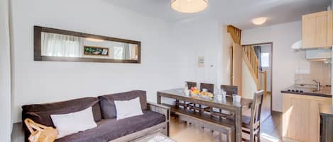 A modern living room with a sofa, coffee table, and dining table. There is a kitchen area with wooden cabinets and a staircase.