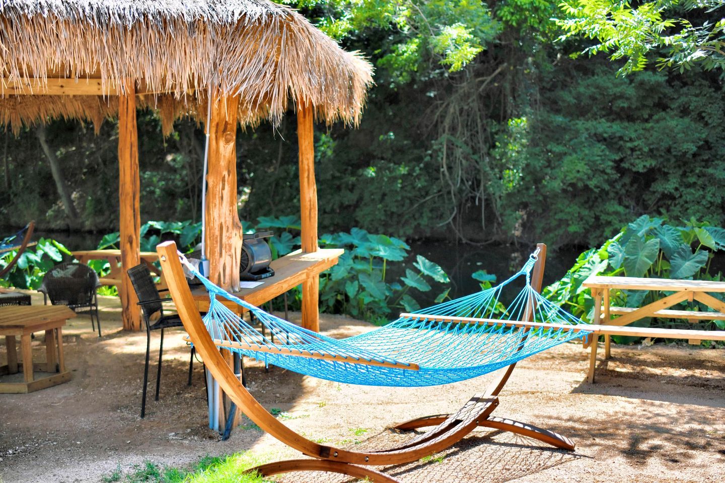 A hammock under a thatched roof structure, surrounded by wooden furniture and greenery.