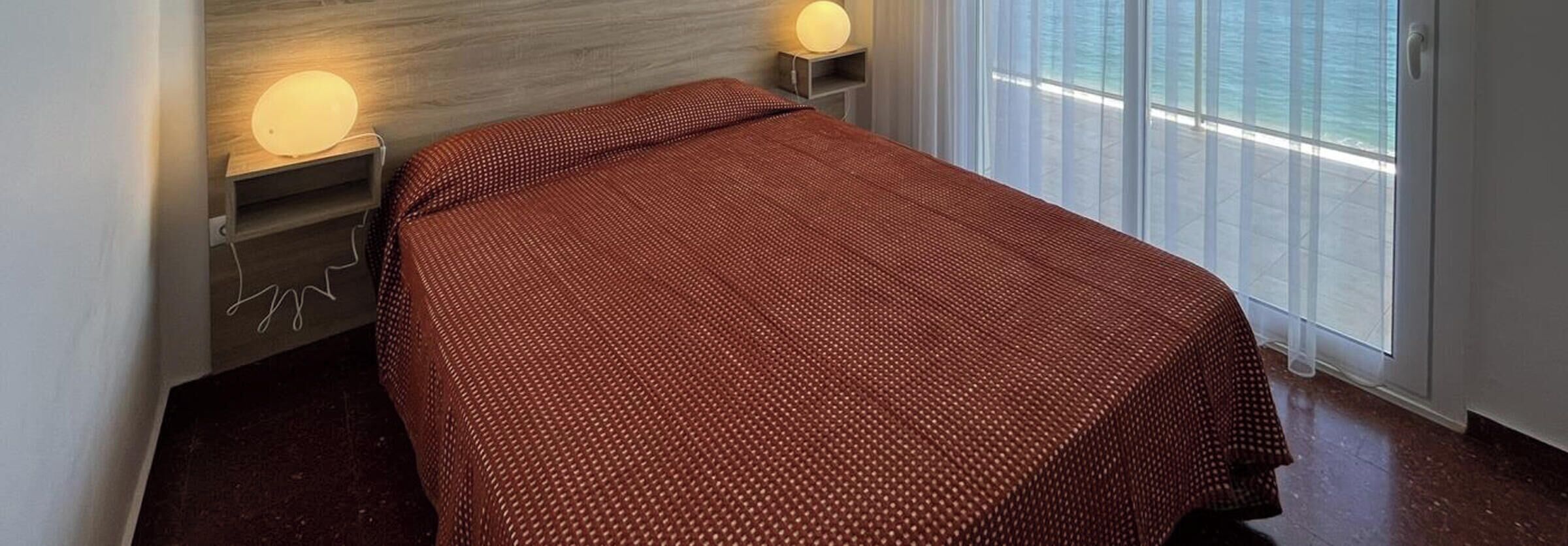 Een enkel bed met een bedovertrek met patroon, twee nachtlampjes, een nachtkastje en een raam met uitzicht.