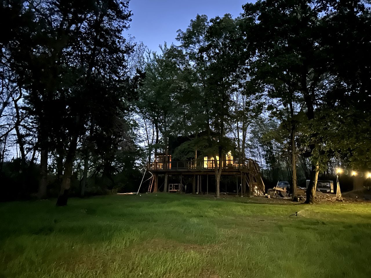 A treehouse structure with a wooden deck and a ladder, surrounded by trees and grass.