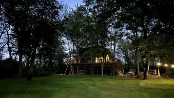 A treehouse structure with a wooden deck and a ladder, surrounded by trees and grass.