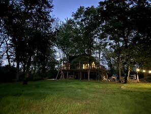 Uma cabana na árvore com um deck de madeira e uma escada, rodeada por árvores e grama.