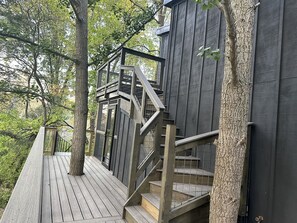 En träveranda med en trappa som leder upp till en byggnad i mörk färg, omgiven av träd.