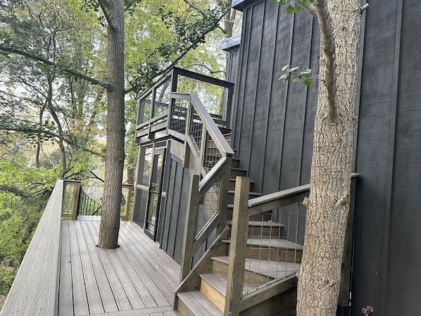 A wooden deck with a staircase leading to a dark-colored building, surrounded by trees.
