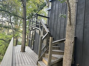 Una terraza de madera con una escalera que lleva a un edificio de color oscuro, rodeado de árboles.