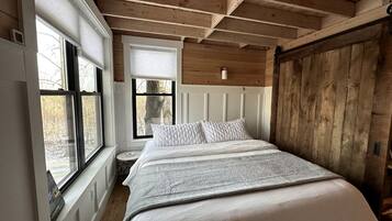 Une chambre avec un grand lit, des poutres apparentes en bois au plafond, une fenêtre avec des stores et une vue sur les arbres à l’extérieur.