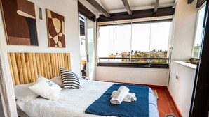 Een slaapkamer met een bed, kussens, een blauwe deken en uitzicht op gebouwen door een raam.