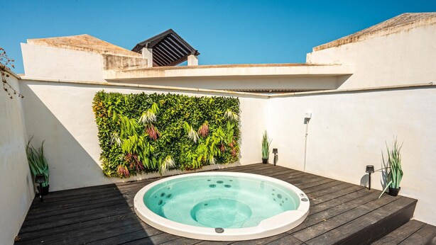 Een hottub op een houten terras, met een verticale tuin aan de muur en een wit gebouw op de achtergrond.