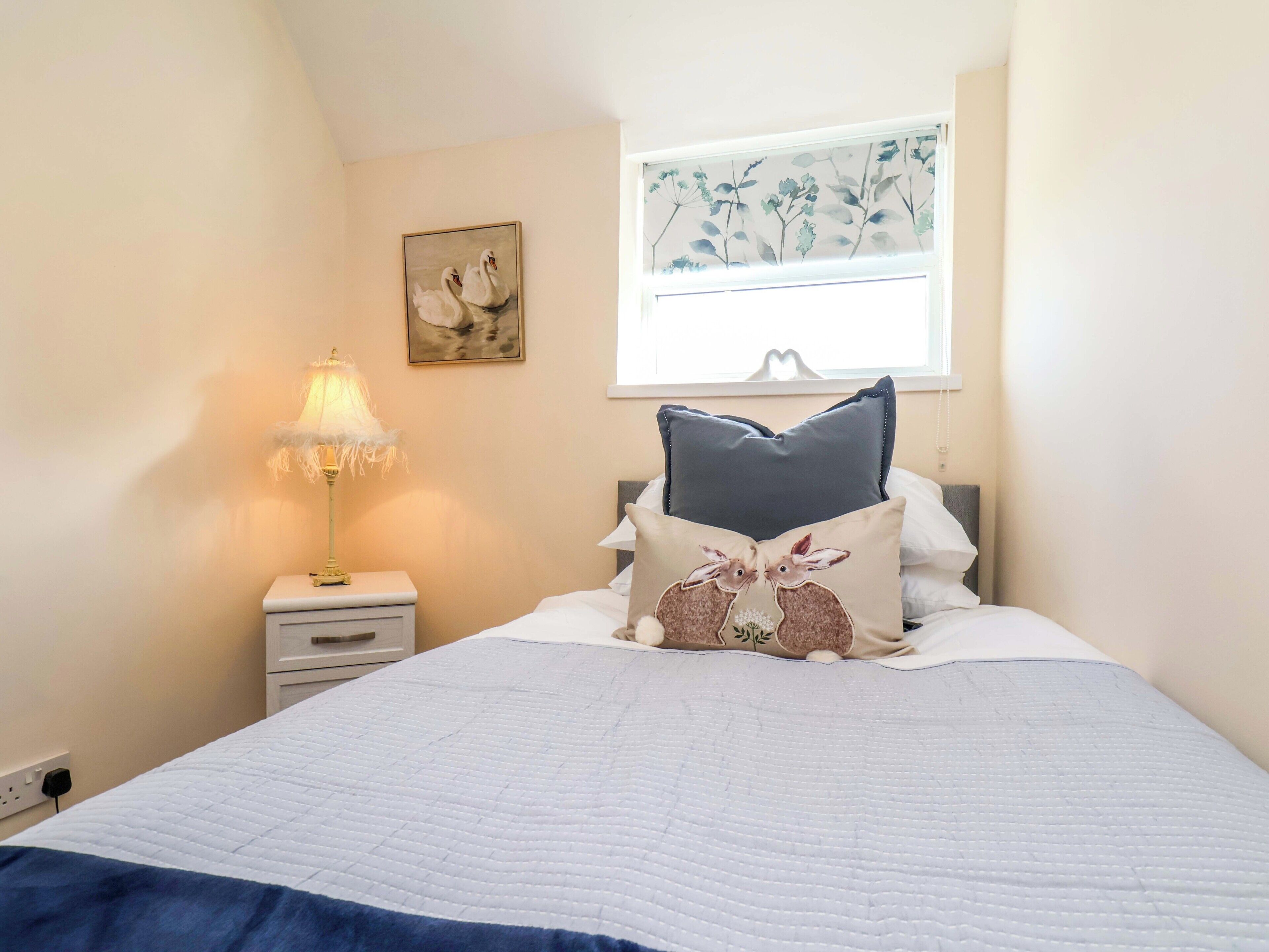 A bedroom with a bed, a nightstand with a lamp, a window with a floral view, and a wall painting.