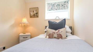 A bedroom with a bed, a nightstand with a lamp, a window with a floral view, and a wall painting.