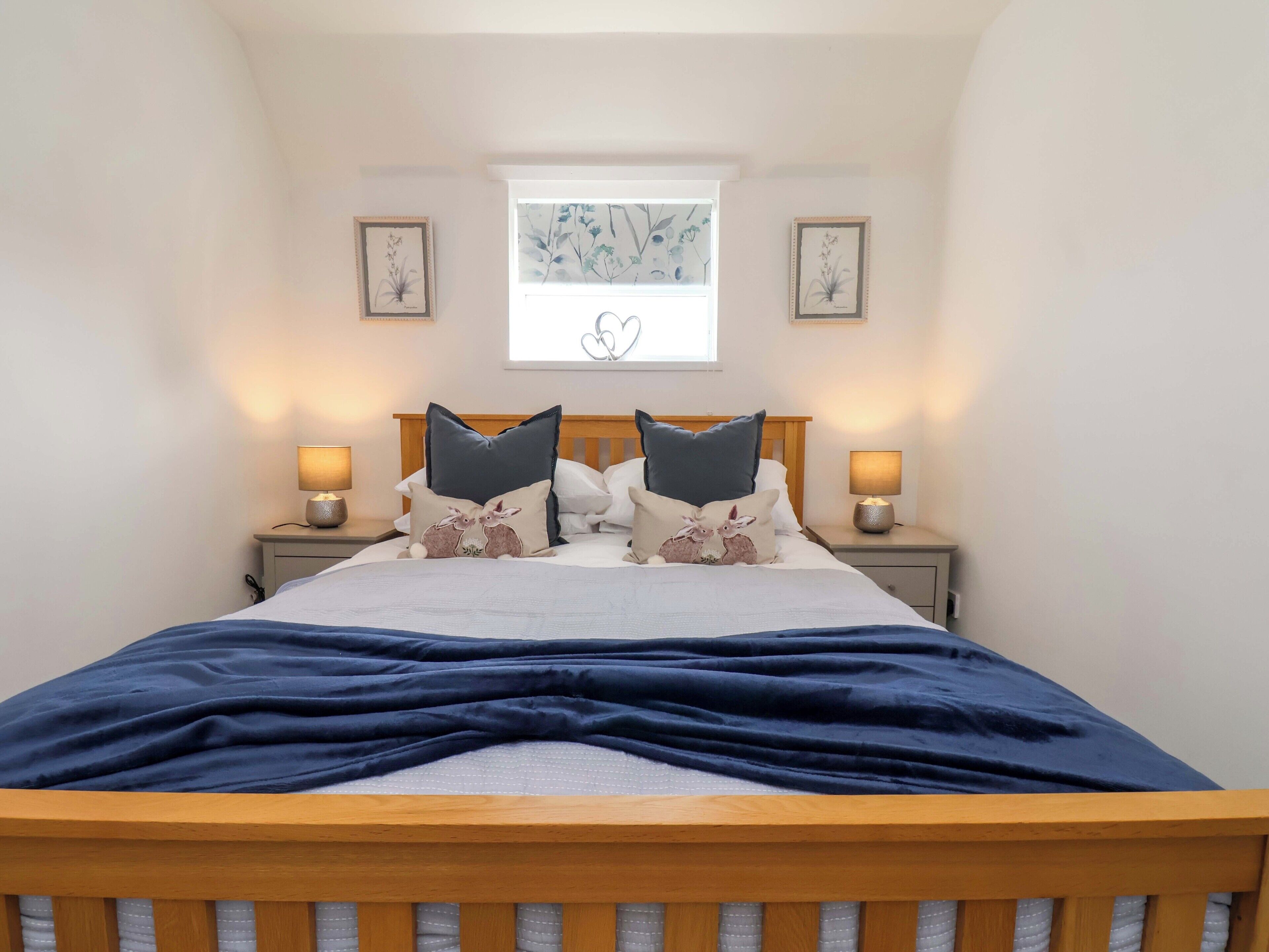 A bedroom with a wooden bed, two bedside tables with lamps, a window with a floral view, and framed botanical prints on the walls.