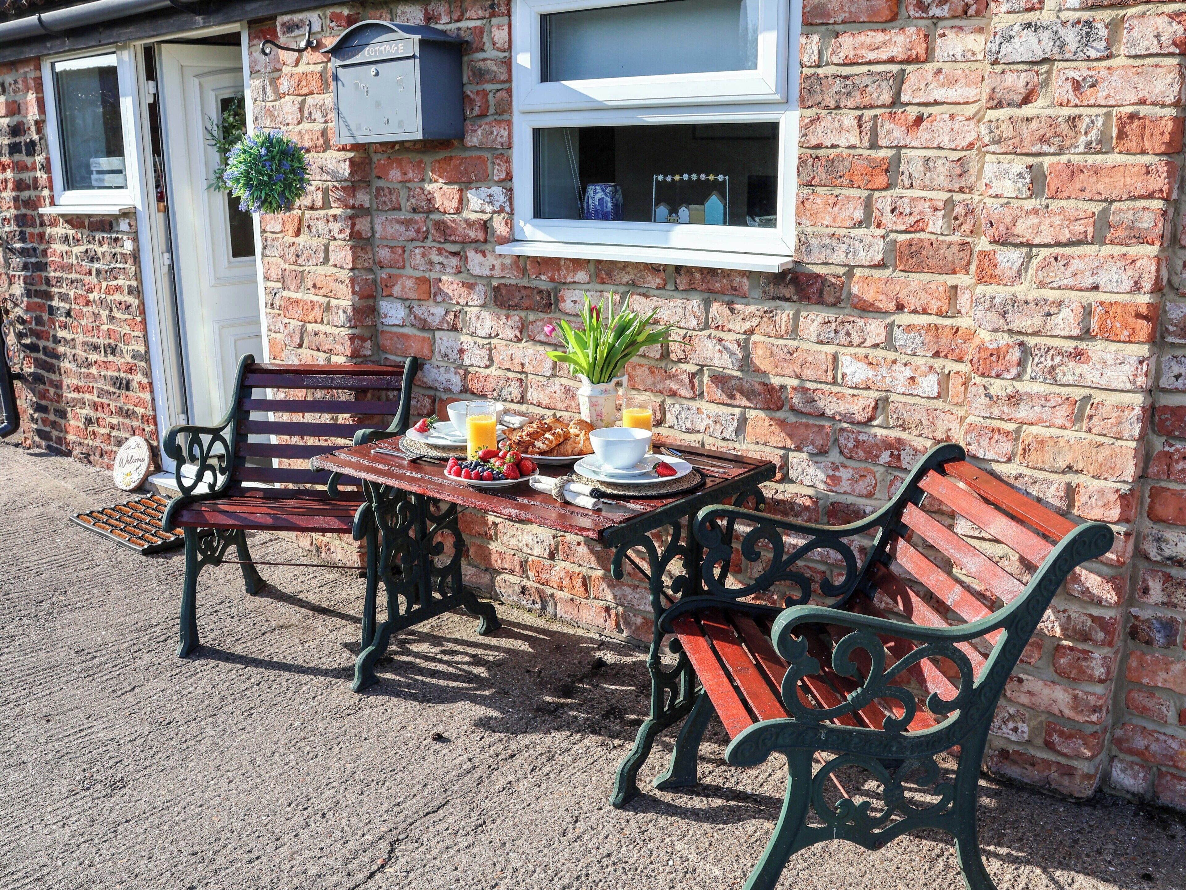 A brick building with a cottage sign, a bench, a table with breakfast, and a window with a plant.