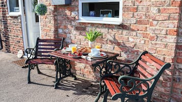 A brick building with a cottage sign, a bench, a table with breakfast, and a window with a plant.
