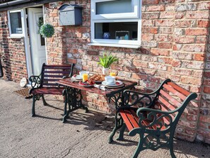 A brick building with a cottage sign, a bench, a table with breakfast, and a window with a plant. - Lamb Cottage (Billingham)