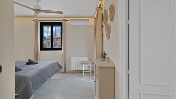 Een slaapkamer met een bed, een plafondventilator, een radiator, een klein tafeltje en een raam met gordijnen.
